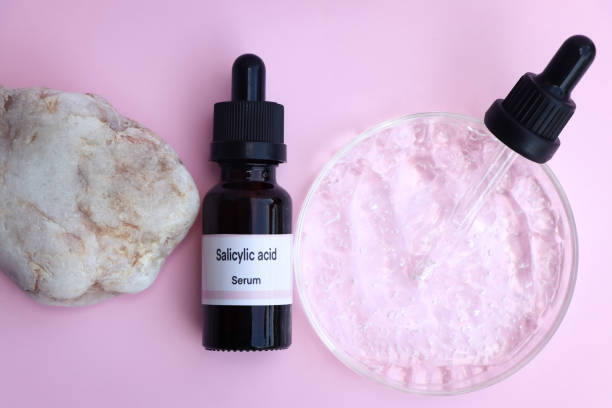 A bottle of salicylic acid serum with a stone and a glass dish containing a clear liquid on a pink background.